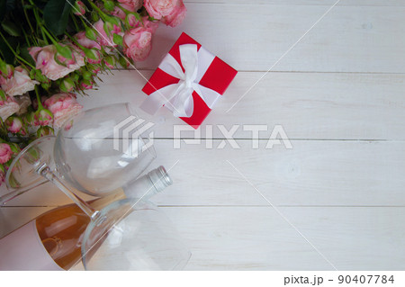 A large bouquet of pink roses, a gift in a red box and a bottle of rose wine with glasses lie on the side on a wooden white background with a place for text. High quality photo 90407784