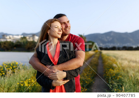 Portrait of lovely middle-aged couple looking aside at copyspace and hugging while standing on grass in summer field. 90407785
