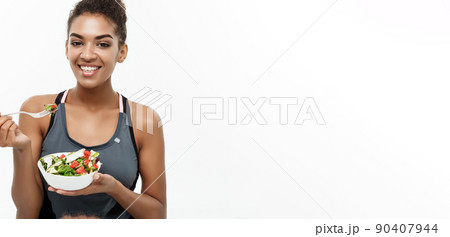 Healthy and Fitness concept - Beautiful American African lady in fitness clothes on diet eating fresh salad. Isolated on white background. Healthy and Fitness concept - Beautiful American African lady in fitness clothes on diet eating fresh salad. Isolated on white background. 90407944