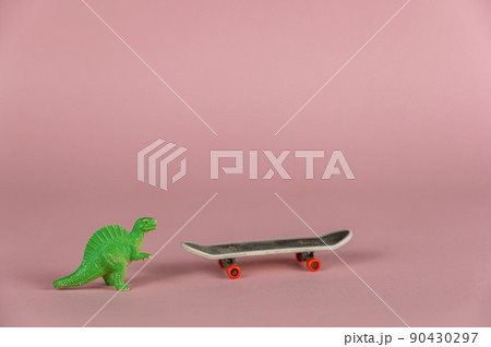 Miniature dinosaur standing next to a mini skateboard. Small green children's toy against a pink background. Close-up. Selective focus. Miniature dinosaur standing next to a mini skateboard. Small green children's toy against a pink background. Close-up. Selective focus. 90430297