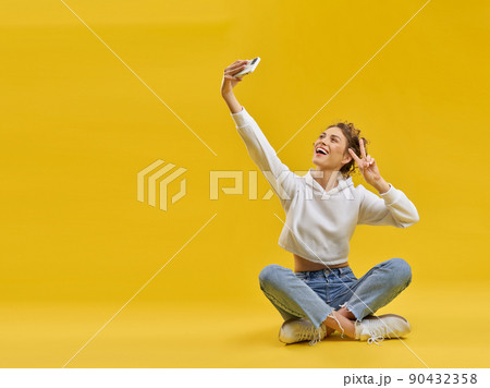 Young girl taking selfie with smartphone, sitting with crossed legs in studio. Front view of smiling female showing victory sign, taking selfie photo, isolated on orange background. Concept of selfie. Young girl taking selfie with smartphone, sitting with crossed legs in studio. Front view of smiling female showing victory sign, taking selfie photo, isolated on orange background. Concept of selfie. 90432358