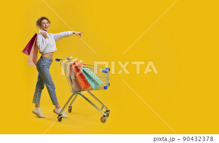 Beautiful female shopaholic pointing away, while shopping in studio. Side view of cute girl with shopping trolley, tiptoeing, looking at camera, isolated on orange background. Concept of shopping. Beautiful female shopaholic pointing away, while shopping in studio. Side view of cute girl with shopping trolley, tiptoeing, looking at camera, isolated on orange background. Concept of shopping. 90432378