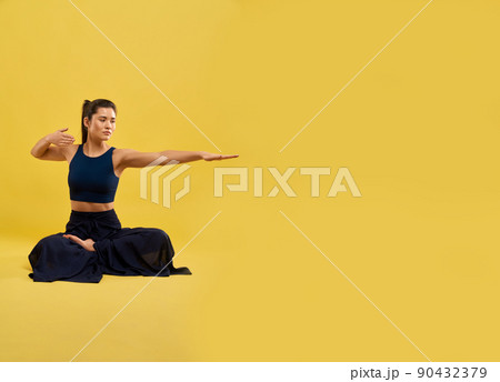 Serious woman sitting in meditation pose, holding arms bent and outstretched in studio. Front view of yogi stretching hand, while sitting in lotus pose, isolated on yellow background. Concept of yoga. 90432379