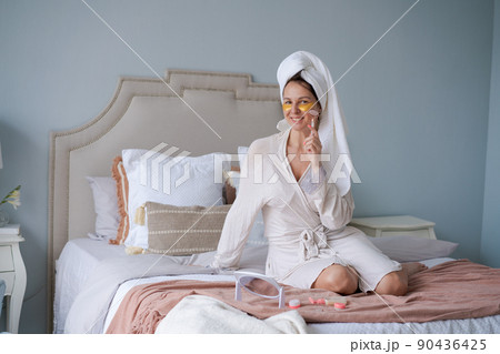 Happy woman sitting in bed, doing facial massage with jade face roller Happy woman sitting in bed, doing facial massage with jade face roller 90436425