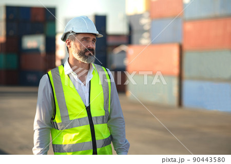 Caucasian male manager in white hard hat helmet and high-visibility vest walking in Container Terminal. 90443580