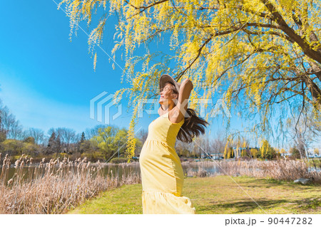 Happy Asian pregnant woman dancing free in nature summer park wearing sun hat and yellow dress. Pregnancy joy and carefree 90447282