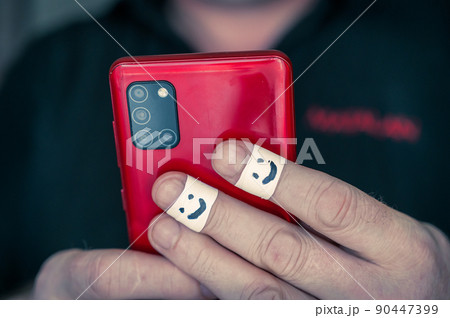 A man is holding a red smartphone. The first phalanges of his index and middle fingers are wrapped in white ribbon. A smiling face is painted on the bandages. Selective focus. 90447399