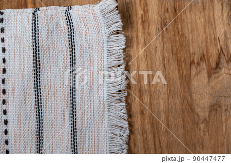 A white kitchen towel lies on the countertop of a wooden table. The fringed towel is fringed and patterned on the rough boards. Top view. Copy space A white kitchen towel lies on the countertop of a wooden table. The fringed towel is fringed and patterned on the rough boards. Top view. Copy space 90447477