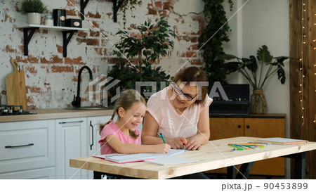 Grandmother Helps to Granddaughter with Homework while Sitting at Table in the Kitchen. Education, Teaching, Learning and People Concept 90453899