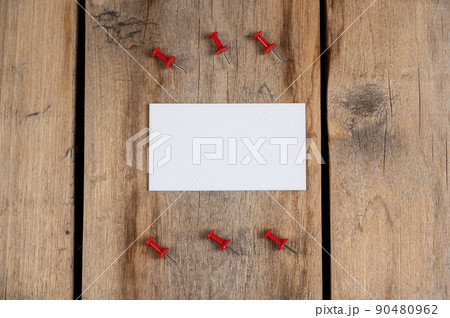 One blank business card and red push pins against a wooden background. A white paper rectangle and six push pins lie on top of old cracked boards. Selective focus. One blank business card and red push pins against a wooden background. A white paper rectangle and six push pins lie on top of old cracked boards. Selective focus. 90480962