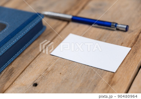 Blue diary, business card and pencil opposite wooden background. Closed hardcover notebook. White blank business card. Metallic silver-blue pencil rests on top of old cracked boards. Selective focus Blue diary, business card and pencil opposite wooden background. Closed hardcover notebook. White blank business card. Metallic silver-blue pencil rests on top of old cracked boards. Selective focus 90480964