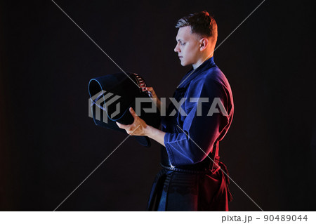 Close up shot, Kendo fighter wearing in an armor, traditional kimono is looking at his helmet while standing against a black studio background. Close up shot, Kendo fighter wearing in an armor, traditional kimono is looking at his helmet while standing against a black studio background. 90489044