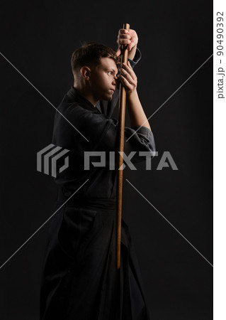 Kendo guru wearing in a traditional japanese kimono is practicing martial art with the shinai bamboo sword against a black studio background. 90490392