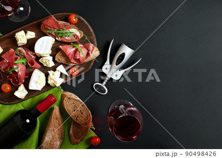 Delicious appetizer to wine - ham, cheese, baguette slices, tomatoes, served on a wooden board, and glass with red wine on black surface 90498426