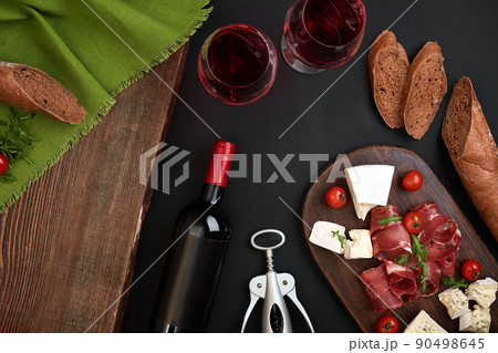 Above overhead view flat lay still life of assortment various cheese and delicatessen with traditional bread and red wine on a old wooden board on black table 90498645