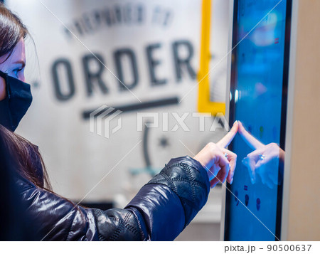 Moscow, Russia -April 3, 2021: Pretty caucasian girl in a mask buys a hot drink at McDonald's. Self service ordering machines inside McDonald's. People walk past a young girl buys coffee using 90500637