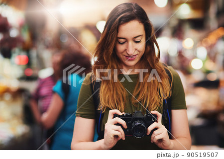 Time to do what tourists do best. Shot of a cheerful young woman holding a camera and looking on the screen while standing at a market outside during the day. 90519507