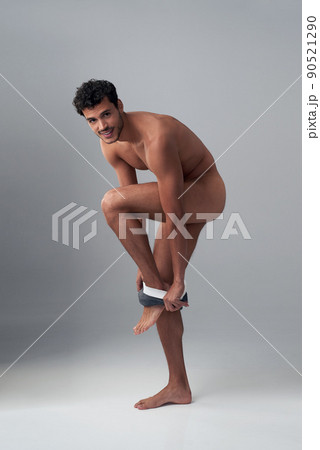 Who needs clothes when you look this good. Studio shot of a handsome young man undressing against a grey background. 90521290