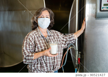 Mature woman in protective mask is holding glass of milk at cow farm 90521427