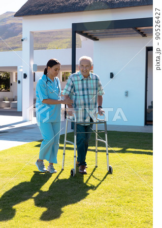 Biracial female doctor assisting smiling caucasian senior man in walking with walker in yard Biracial female doctor assisting smiling caucasian senior man in walking with walker in yard 90526427