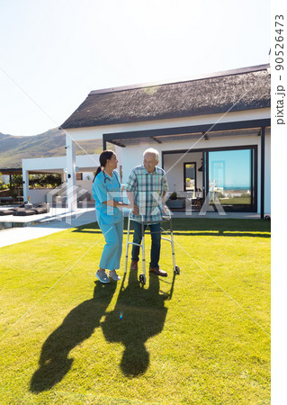 Smiling biracial female doctor assisting caucasian senior man in walking with walker in yard Smiling biracial female doctor assisting caucasian senior man in walking with walker in yard 90526473