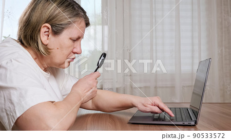 Old woman uses magnifying glass for typing on keyboard Old woman uses magnifying glass for typing on keyboard 90533572