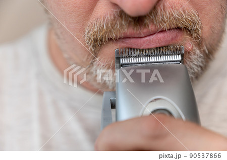 A mature man shaving. The bottom of a man's face. Man cuts facial hair with a hair clipper. A male with stubble. Gray hair on his beard and mustache. Close-up. Selective focus. 90537866