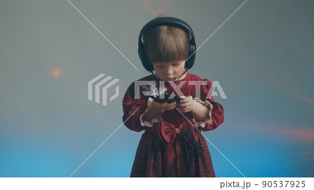 Cute little girl in big headphones and a beautiful vintage dress with lace is listening to music on her phone. There is a color music in the background. The concept of music mania and audiophilia 90537925