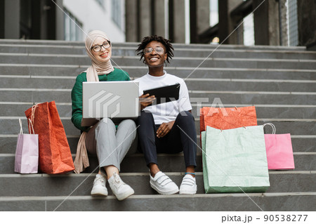 Best friends women making online purchases using a tablet and laptop sitting on steps of the mall 90538277
