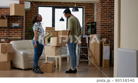 Husband and wife moving in new home with carton boxes, using furniture to start new beginnings together in living room. Holding packages in rented property on loan for relocation. 90538540