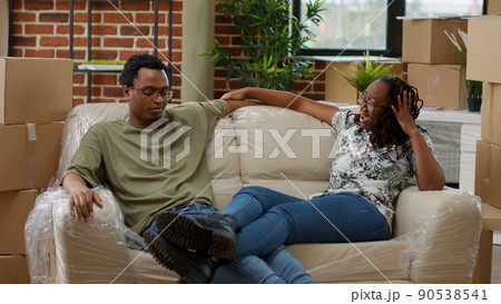 Husband and wife unpacking home decor and relaxing on sofa in new apartment property, taking break after moving in rented flat. Enjoying relocation as homeowners. Handheld shot. 90538541