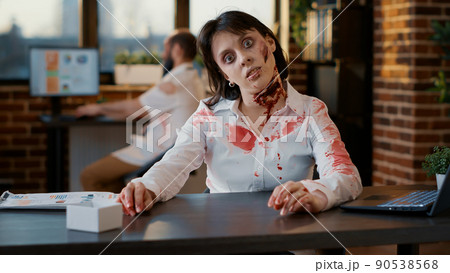 Ugly spooky looking walking dead person sitting in office while waving at camera. Bizarre brain dead monster with bloody and deep scars and wounds sitting in workspace while waving hand at camera. Ugly spooky looking walking dead person sitting in office while waving at camera. Bizarre brain dead monster with bloody and deep scars and wounds sitting in workspace while waving hand at camera. 90538568