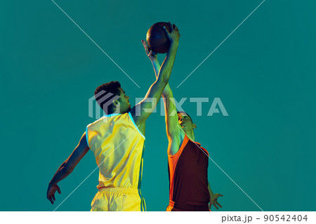 Portrait of two young men, professional basketball players in motion, training isolated over blue background in neon. Cathcing ball in a jump. 90542404