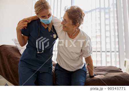 Female doctor in mask helping patient to get up from bed in the ward at hospital Female doctor in mask helping patient to get up from bed in the ward at hospital 90543771