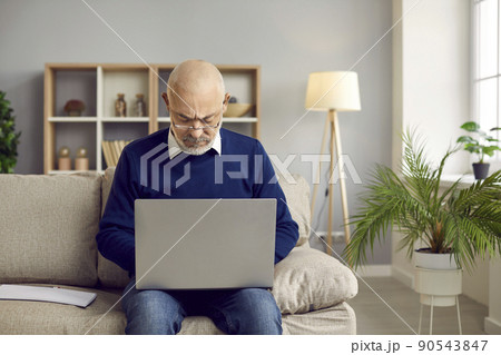 Senior man in glasses sitting on the couch at home and working on his laptop computer 90543847