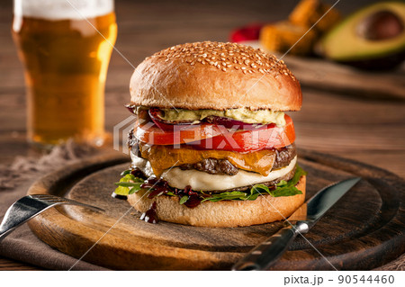 Cheeseburger, made from rye bun with tomato slice and melted cheese on a roasted beef, and lettuce leaf on a wooden table, with a glass of beer 90544460