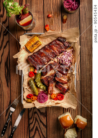 Grilled pork ribs served with grilled corn, salat, bbq sauce, salt pepper and cucumber on parchment paper on a wooden table. Top View 90545434