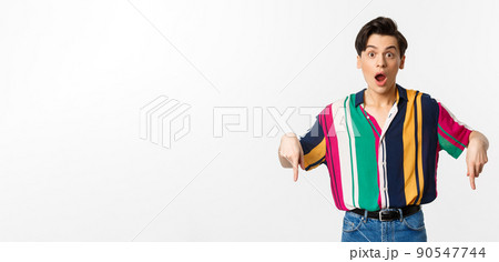 Image of amazed and surprised man showing logo, pointing fingers down and staring with excitement at camera, standing over white background 90547744