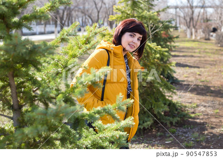 A brunette woman in a yellow jacket peeks out from behind a tree on a walk 90547853