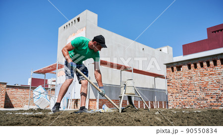 Full length of male worker shoveling sand-cement mix while working at construction site. Man in shorts preparing floor screed material while standing outdoors near concrete screed mixer machine. 90550834