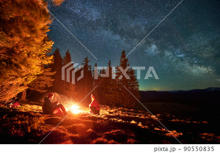 Pair of tourists spending evening time near bonfire on the middle of glade under open fullstar sky with Milky way in the mountains. Tent, trees, mountain peaks on the background. 90550835