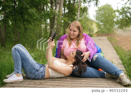 A young brunette woman lies on her girlfriend's lap and shows a video on her smartphone. Two lesbian girls are relaxing outdoors. Rainbow LGBT Flag on girlfriend's shoulder A young brunette woman lies on her girlfriend's lap and shows a video on her smartphone. Two lesbian girls are relaxing outdoors. Rainbow LGBT Flag on girlfriend's shoulder 90552203