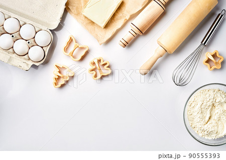 Bowl with wheat flour, rolling pin, whisk, eggs, butter, cookie cutters. Top view on a white table with a copy space Bowl with wheat flour, rolling pin, whisk, eggs, butter, cookie cutters. Top view on a white table with a copy space 90555393