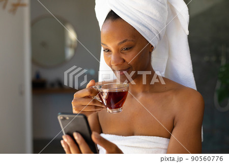 African american woman in a bathrobe holding a tea cup using smartphone at home 90560776