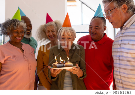Happy multiracial friends wearing party hats looking at senior woman blowing birthday candles 90561262