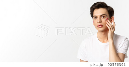 People, lgbtq community and lifestyle concept. Beautiful young gay man applying glitter under eyes for pride party, standing over white background People, lgbtq community and lifestyle concept. Beautiful young gay man applying glitter under eyes for pride party, standing over white background 90576391