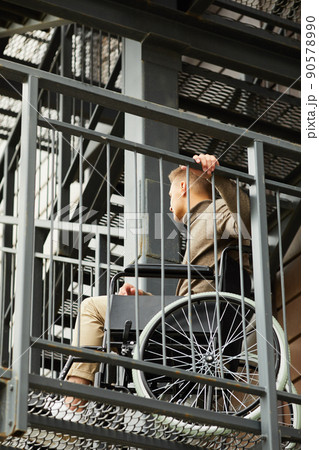 Young handicapped man in brown turtleneck sitting in wheelchair and holding metal railing while trying to go up stairs Young handicapped man in brown turtleneck sitting in wheelchair and holding metal railing while trying to go up stairs 90578990