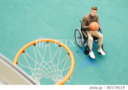 Serious young handicapped man sitting in wheelchair and playing basketball on sports ground Serious young handicapped man sitting in wheelchair and playing basketball on sports ground 90579019