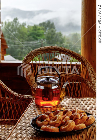 Hot fruit tea in a glass teapot and cookies stands on the edge of a rattan table. Hot fruit tea in a glass teapot and cookies stands on the edge of a rattan table. 90579732