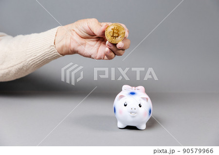 Wrinkled hand of an elderly woman holding a bitcoin near a piggy bank. Gray background. The concept of savings and cryptocurrency Wrinkled hand of an elderly woman holding a bitcoin near a piggy bank. Gray background. The concept of savings and cryptocurrency 90579966